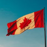 Canadian flag blowing in the breeze on a flagpole, in front of a blue sky.