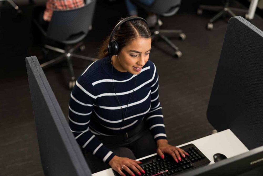 Woman sat at a computer in an IELTS testing booth with headphones on, with others in the background.